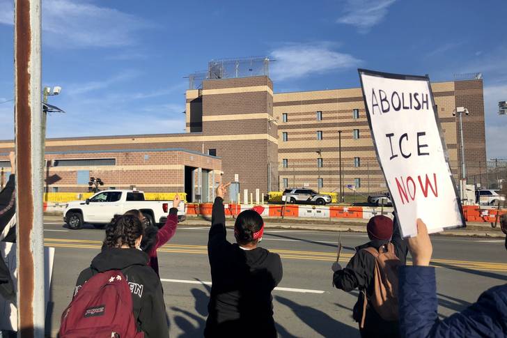 Protesters, including one carrying an "Abolish ICE Now" sign, stand across the street from the brown and beige jail